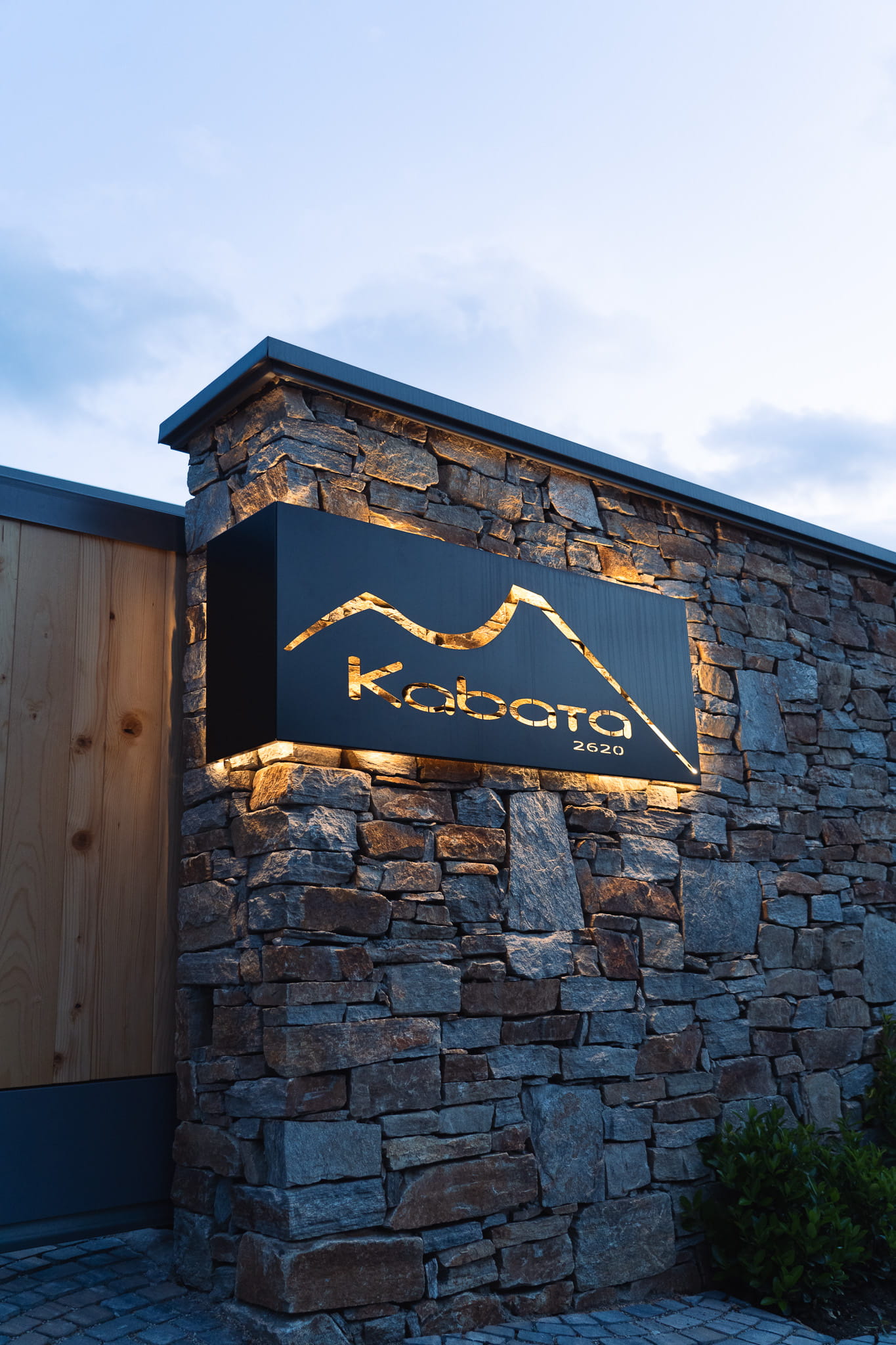 Illuminated black sign with mountain outline and text 'Kabata 2620' on stone wall at dusk.