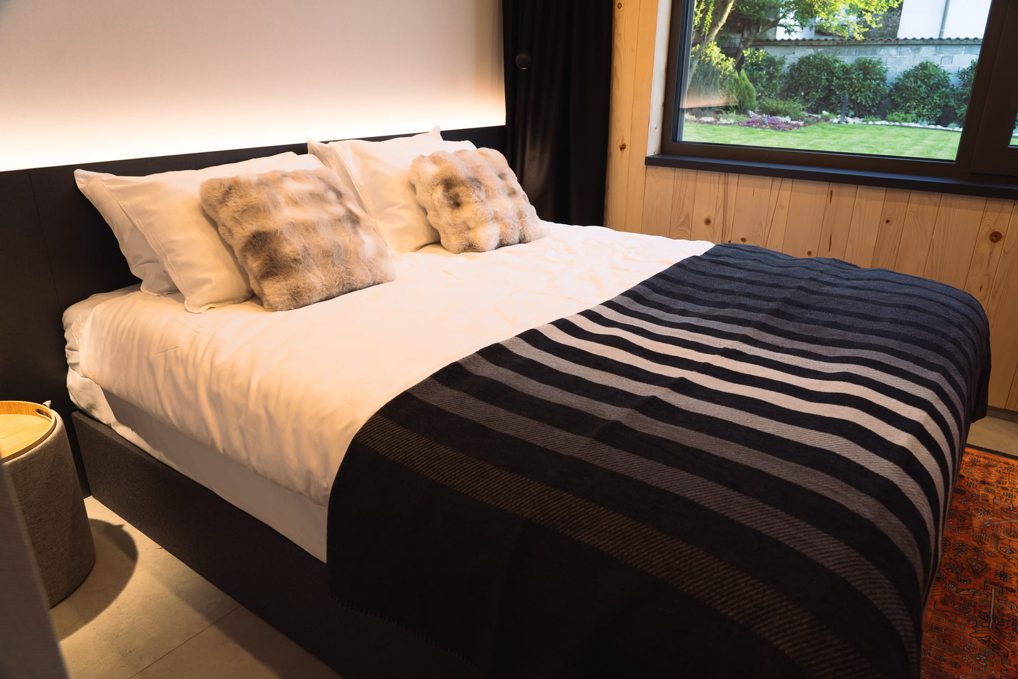 Cozy bed with white sheets, fur pillows, and striped blanket next to a window with garden view.