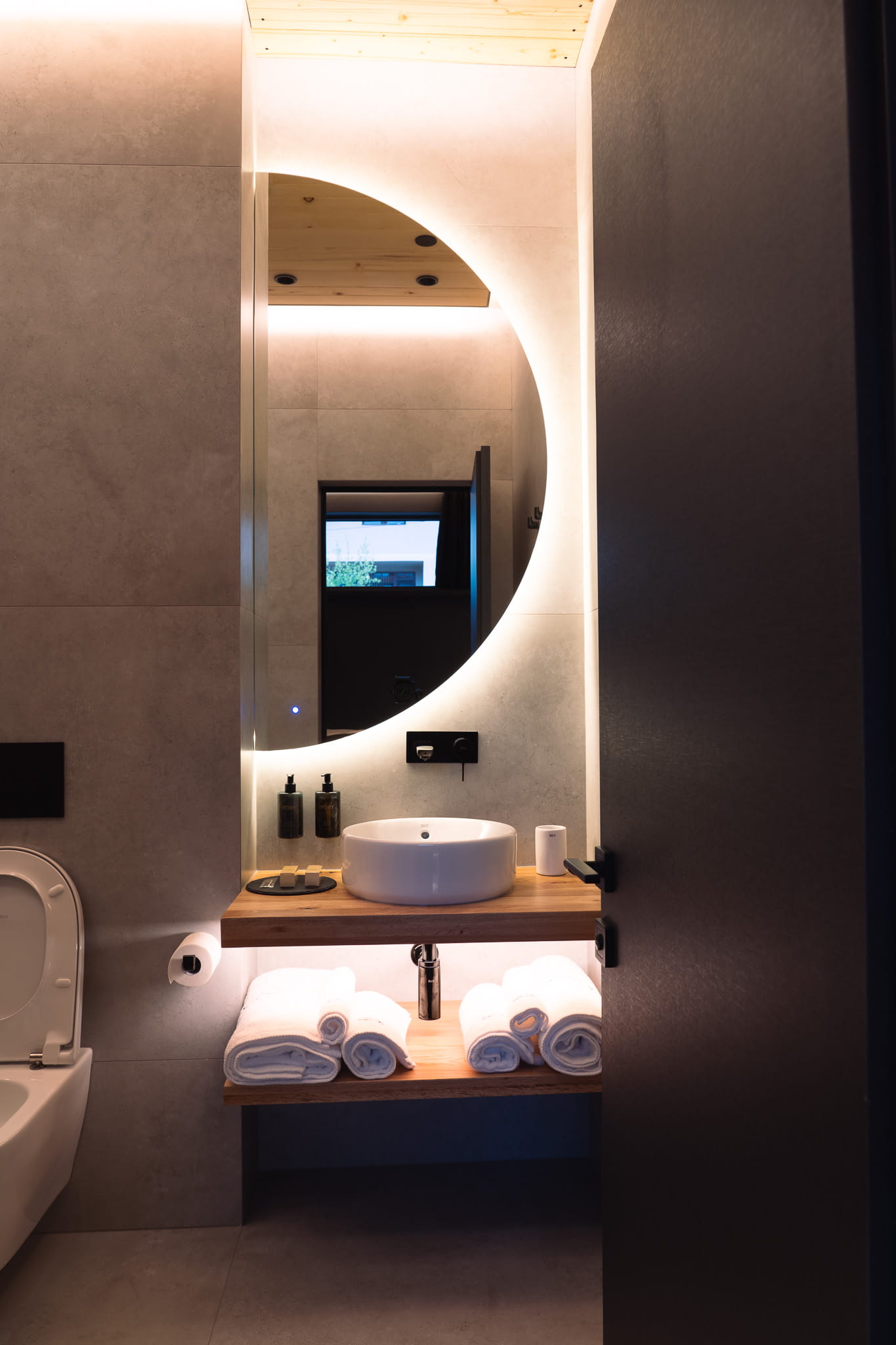 Modern bathroom with round illuminated mirror, countertop sink, toiletries, and rolled white towels on wooden shelves.