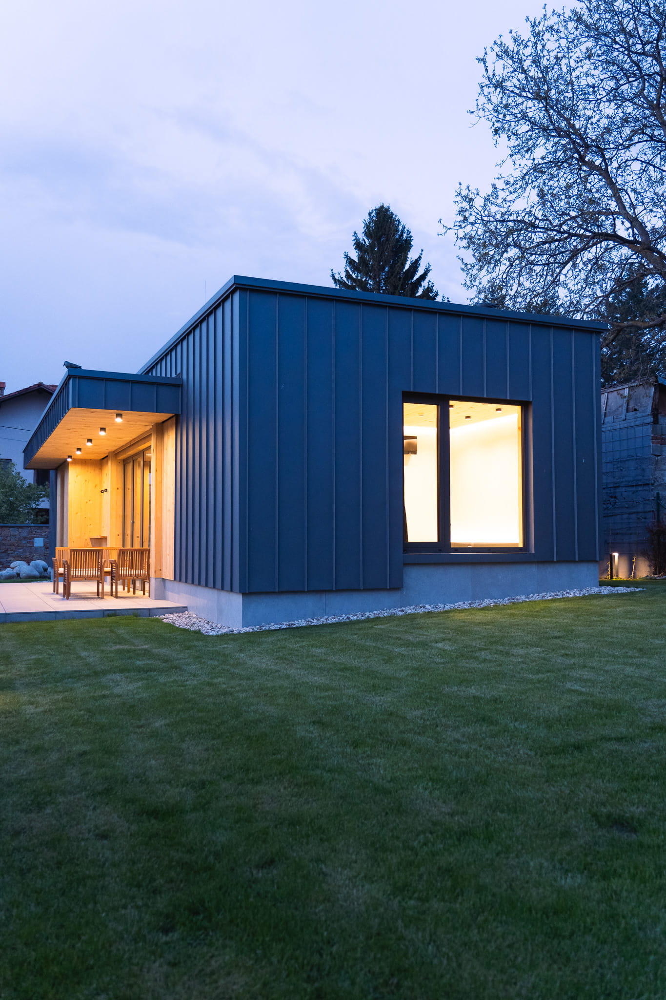 Modern single-story house with dark vertical metal siding, a large window, and a patio with wooden chairs on a green lawn.