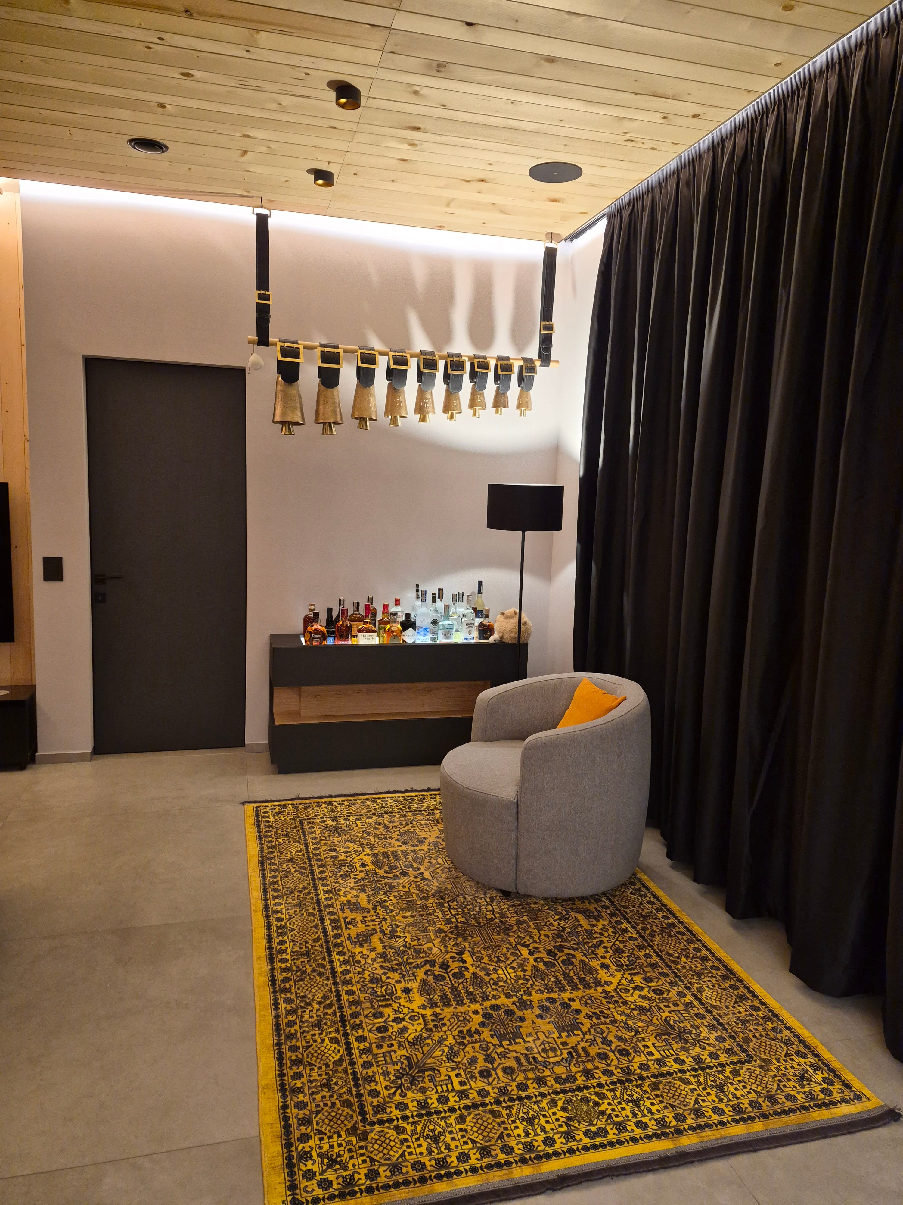 Modern living room corner with a gray round armchair featuring an orange cushion, a yellow patterned rug, a black bar cabinet with various liquor bottles, a black floor lamp, and large hanging decorative bells above.
