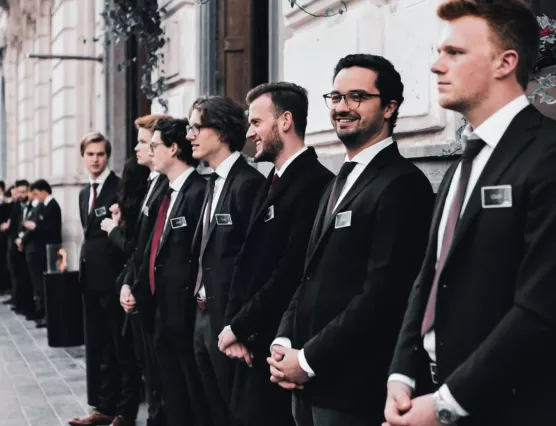 Line of men dressed in formal suits standing outdoors against a building wall, some smiling and wearing name tags.