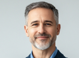 Smiling middle-aged man with gray hair and beard wearing a dark blazer and blue shirt.