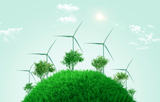 Wind turbines with trees on a green grassy hill under a bright sky with sun and clouds.