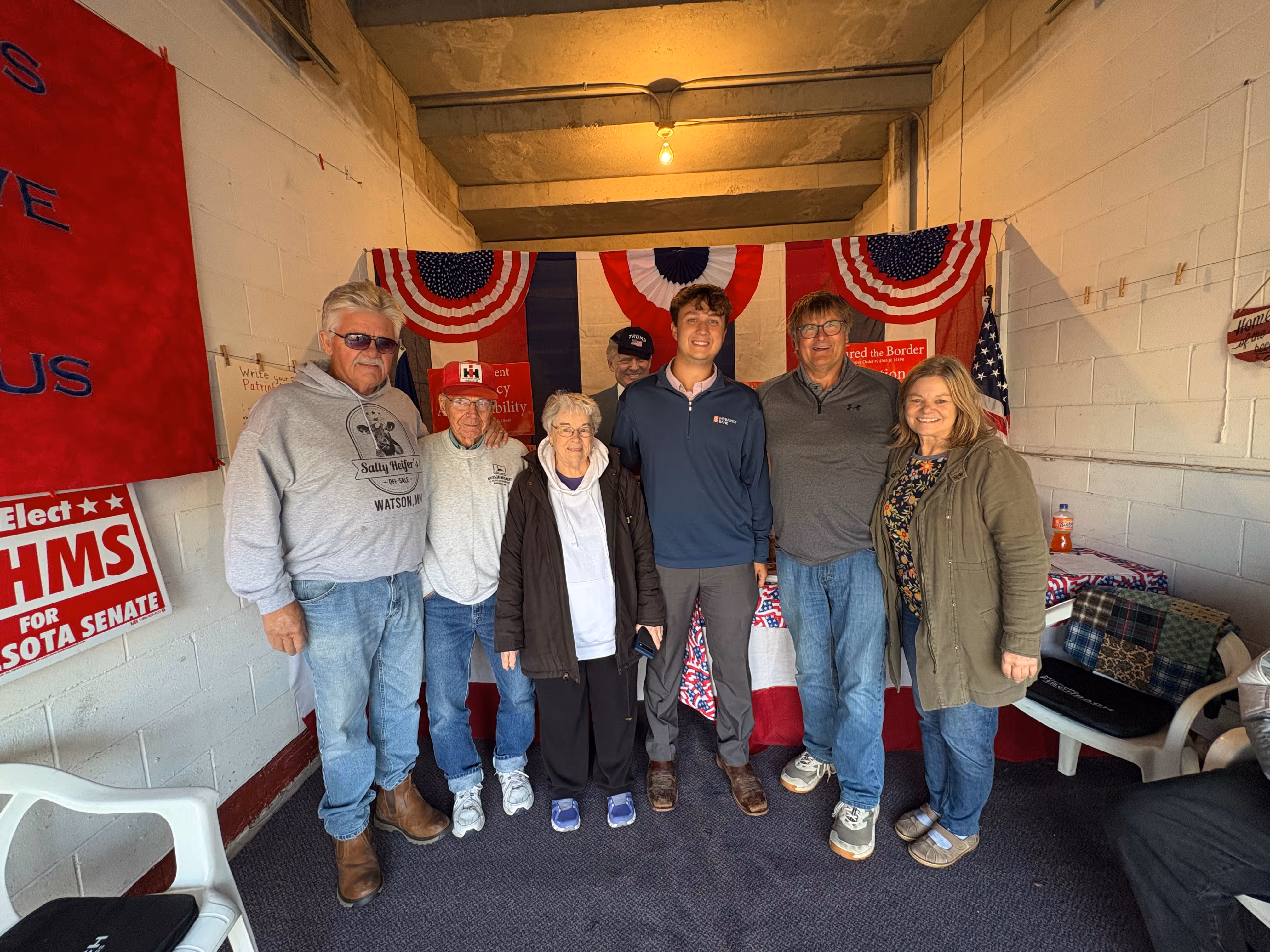 Group of six people standing indoors in front of patriotic red, white, and blue decorations with political campaign signs on the wall.