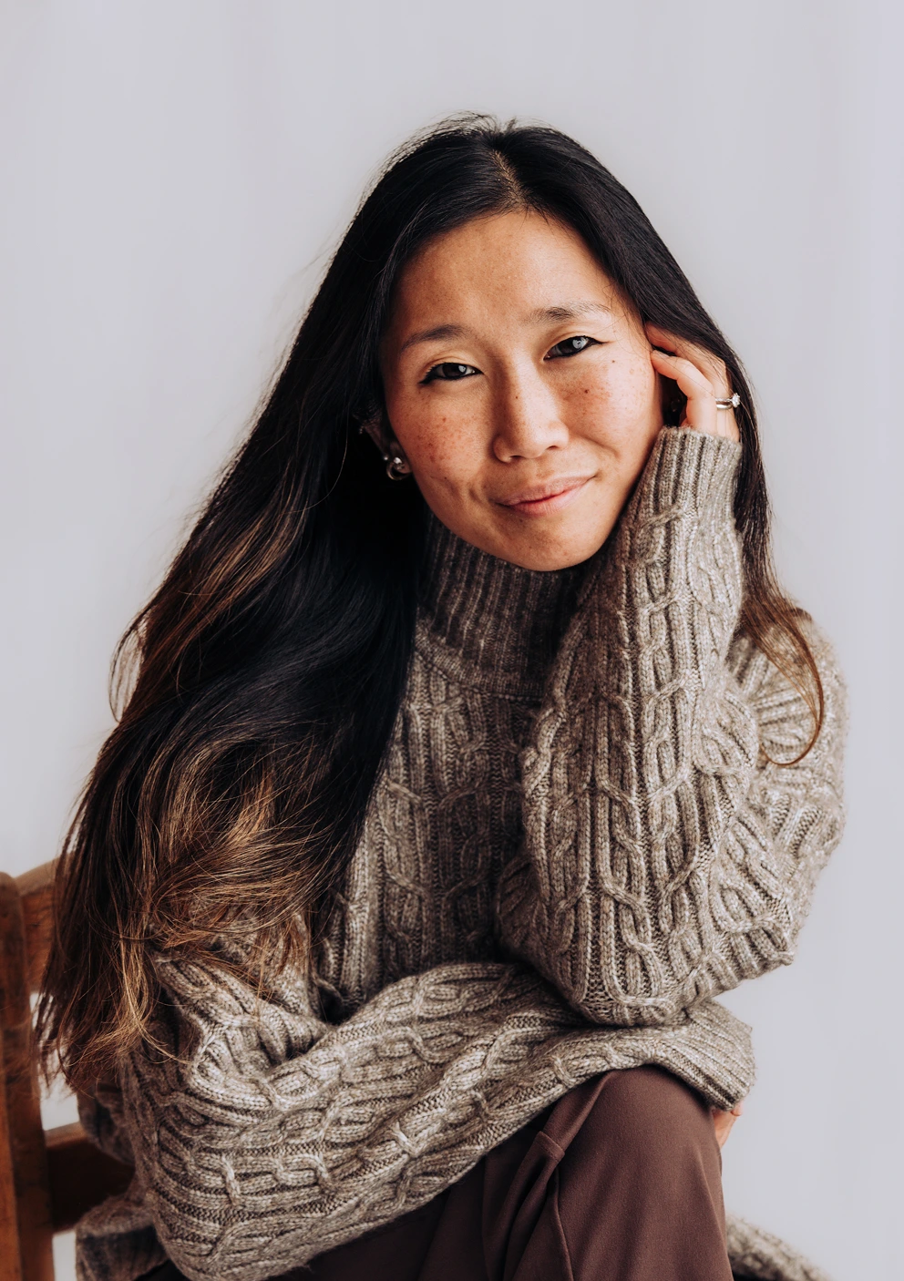 Founder Rachel Forbes wearing a gray cable-knit sweater, sitting and gently touching her face while smiling softly.