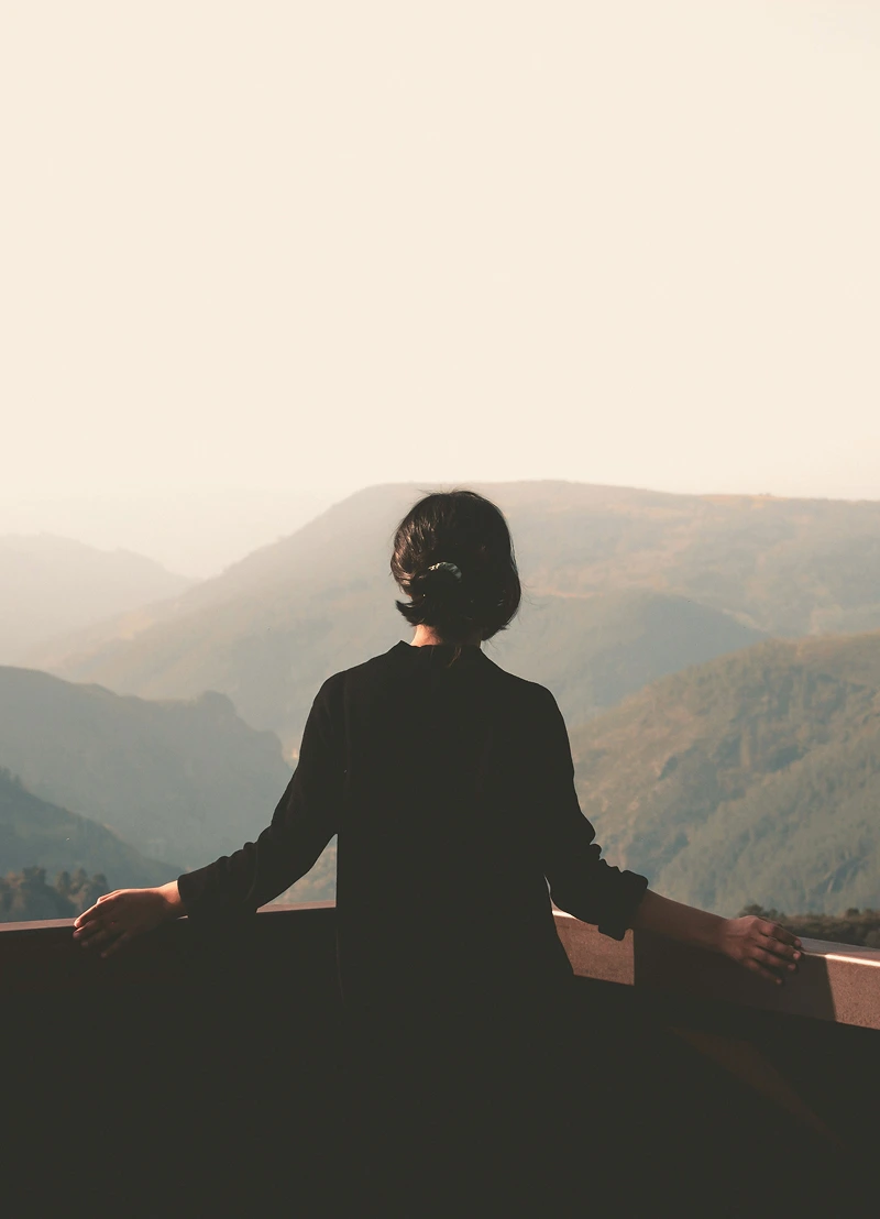 Woman in black clothing standing with arms resting on a railing, overlooking misty mountains in the distance.