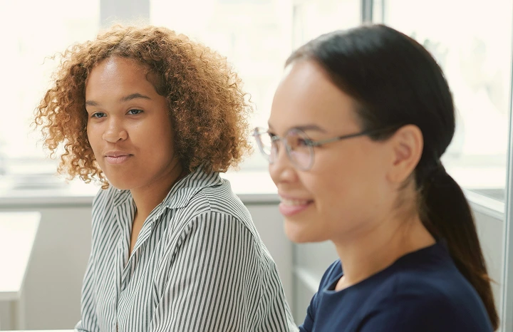 Two women sitting side by side in a bright office, one with curly hair in a striped shirt and the other with glasses and a ponytail.