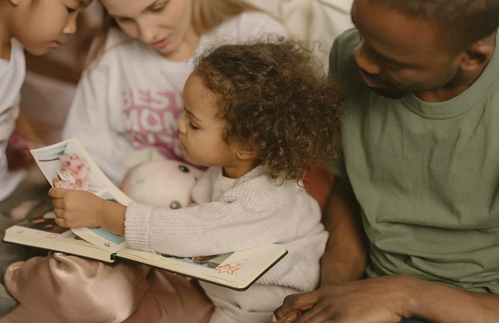 A young child with curly hair reading a book while sitting between an adult woman and an adult man.