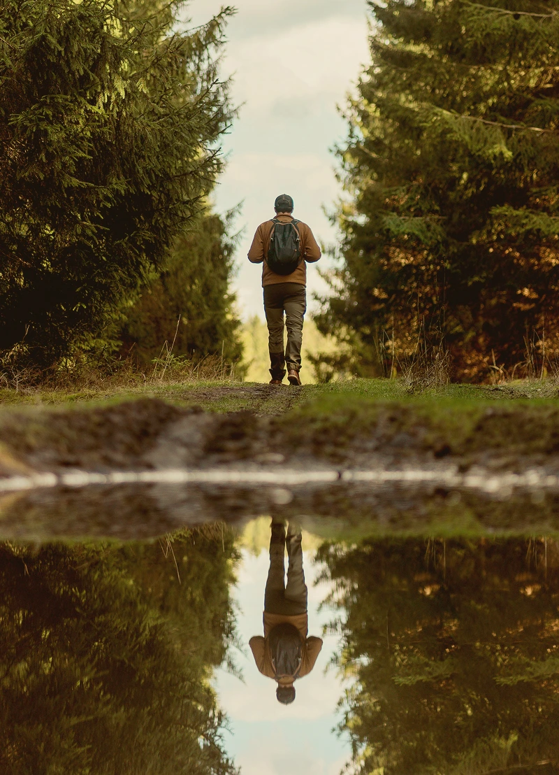 Person wearing a backpack walking on a forest path with their reflection visible in a water puddle.