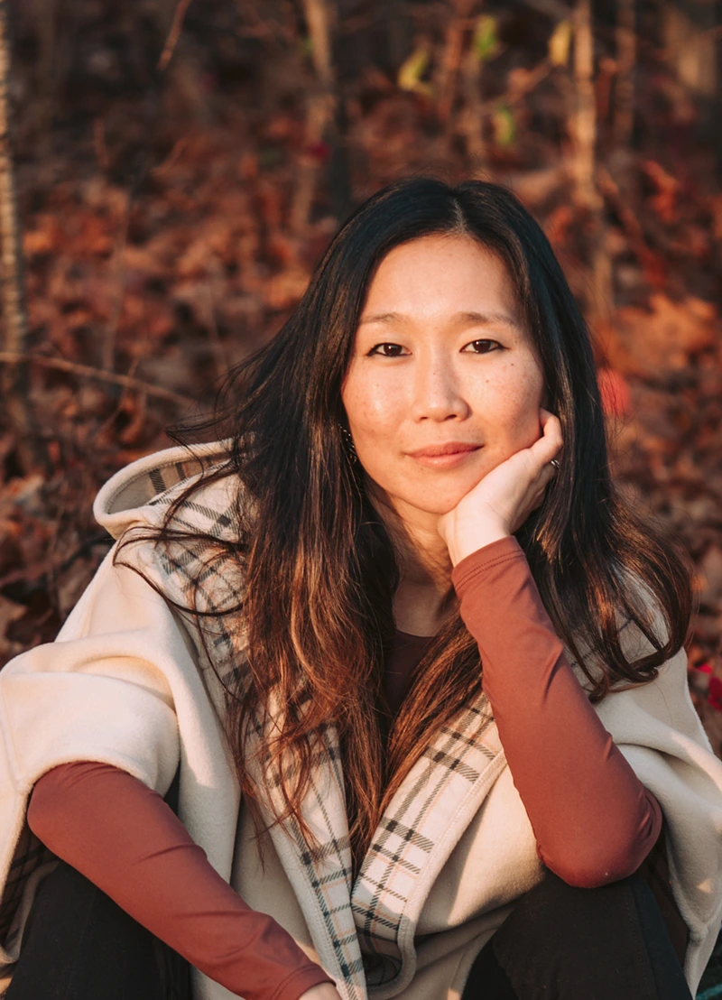 Founder Rachel Forbes wearing a beige plaid-lined coat and maroon shirt sitting outdoors with hand resting on face.