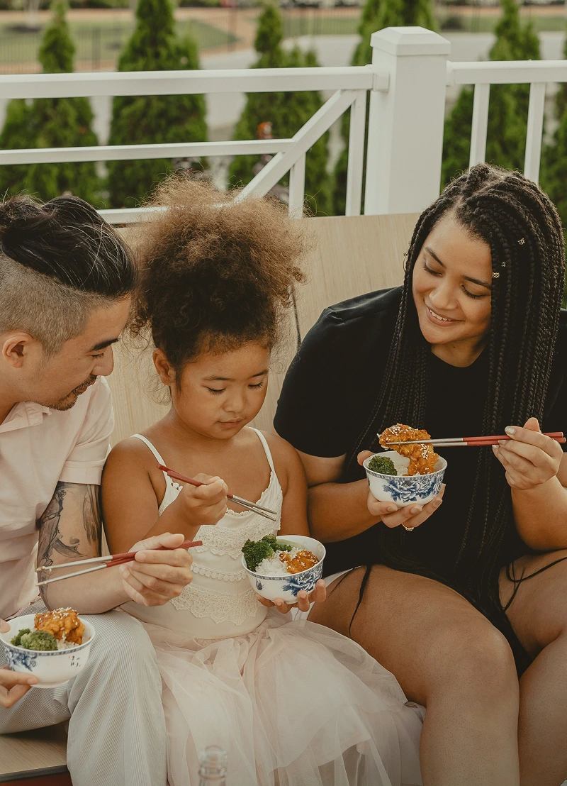 Family of three sitting outdoors eating rice bowls with chopsticks and smiling.