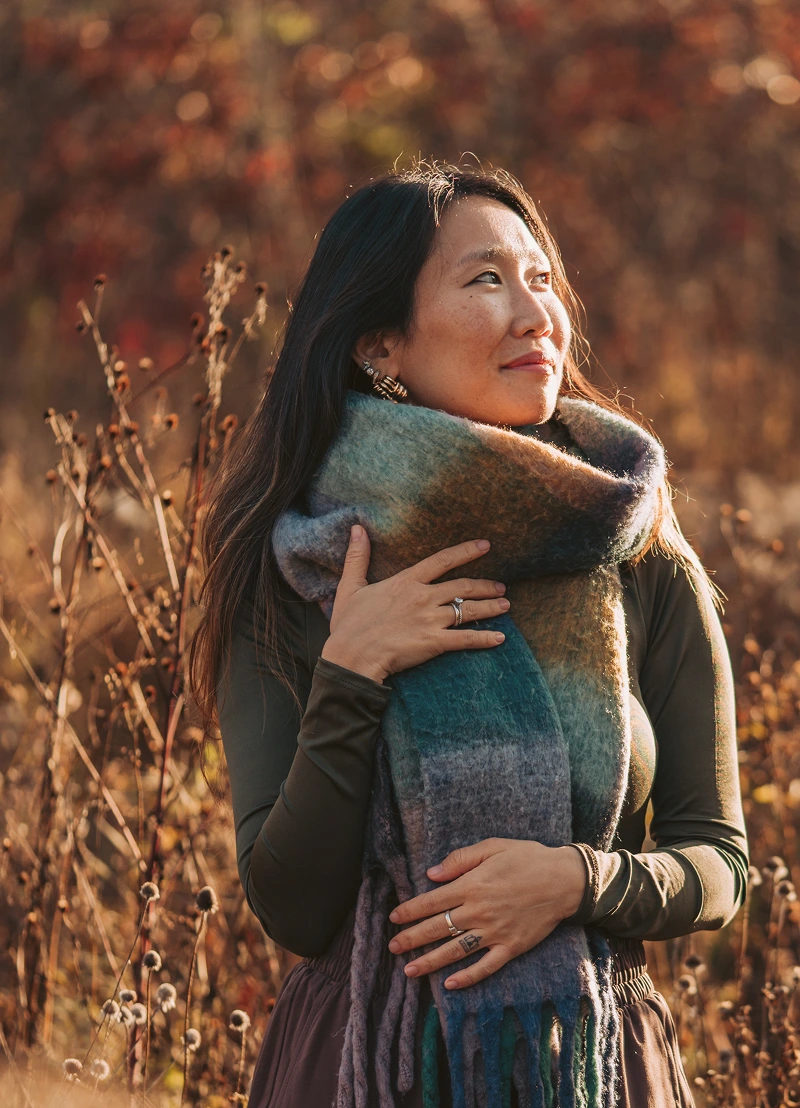 Founder Rachel Forbes standing outdoors in autumn, wrapped in a large multicolored scarf, looking to the side with dry plants in the background.