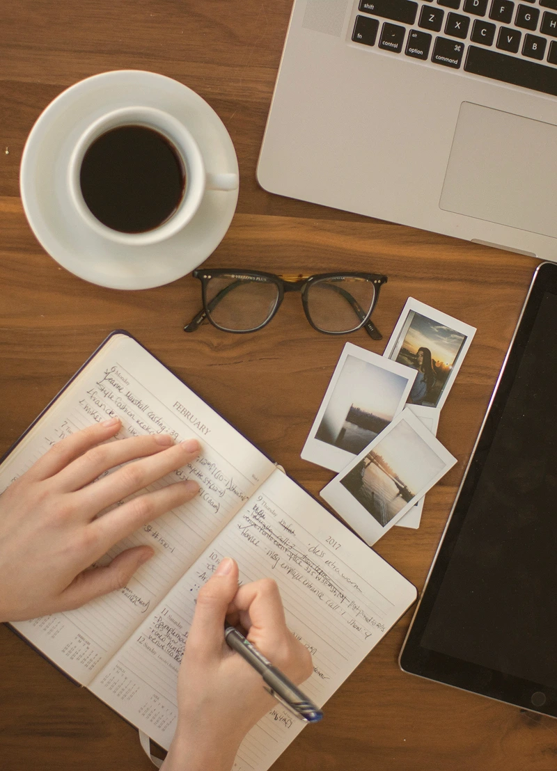 Overhead view of hands writing in a planner with a pen on a wooden table, next to a cup of coffee, eyeglasses, three instant photos, a laptop, and a tablet.
