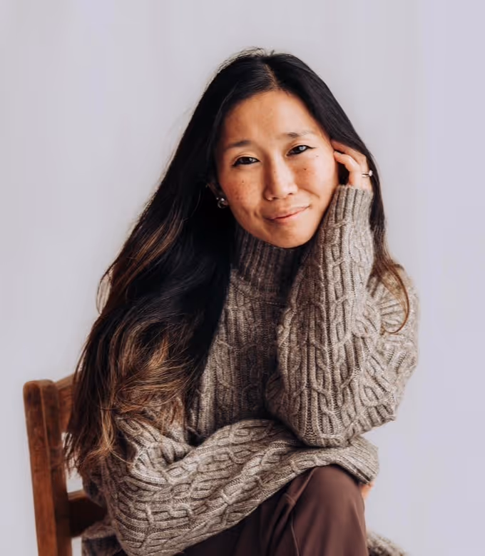Founder Rachel Forbes in a black ribbed top leaning on the back of a wooden chair, smiling gently.
