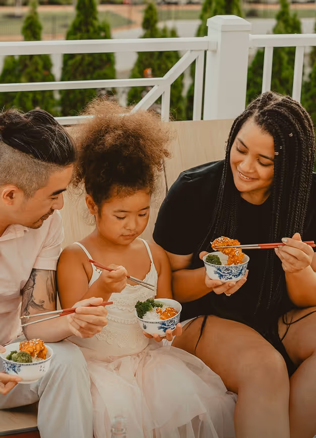 Three people sitting outdoors eating from bowls with chopsticks, smiling and engaging together.