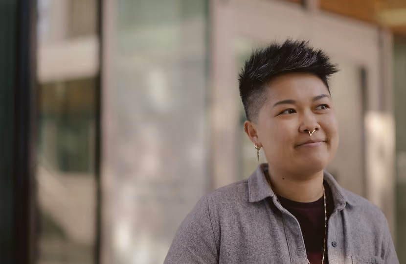 Person with short, spiked black hair wearing a gray button-up shirt over a dark top, with a septum ring and earrings, looking slightly to the side with a thoughtful expression.