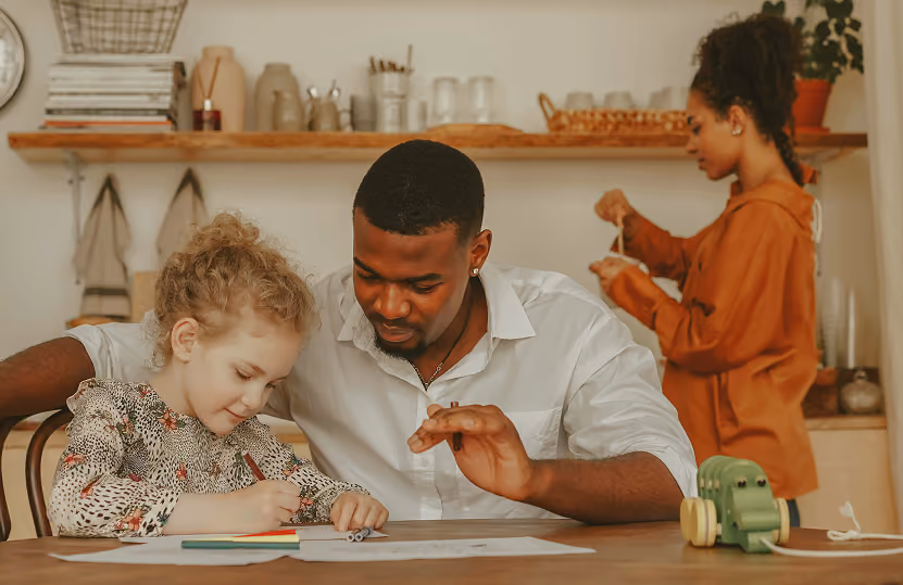 A man in a white shirt helps a young girl with curly hair as she draws with colored pencils at a table, while a woman in an orange shirt stands in the background engaged in an activity.