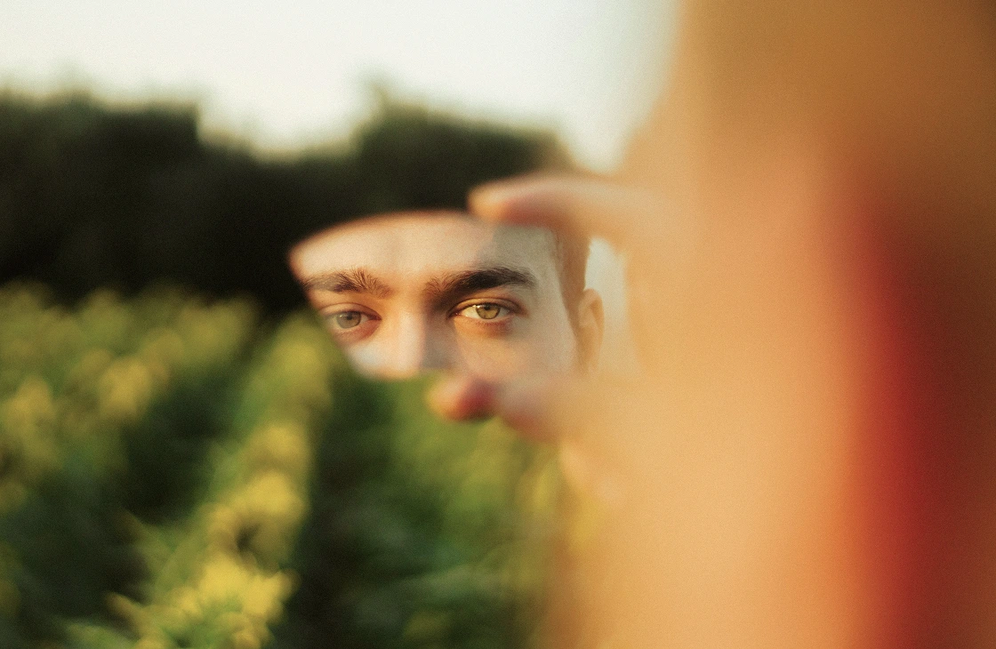 Close-up of a person's eyes reflected in a small broken mirror held outdoors with blurred greenery in the background.