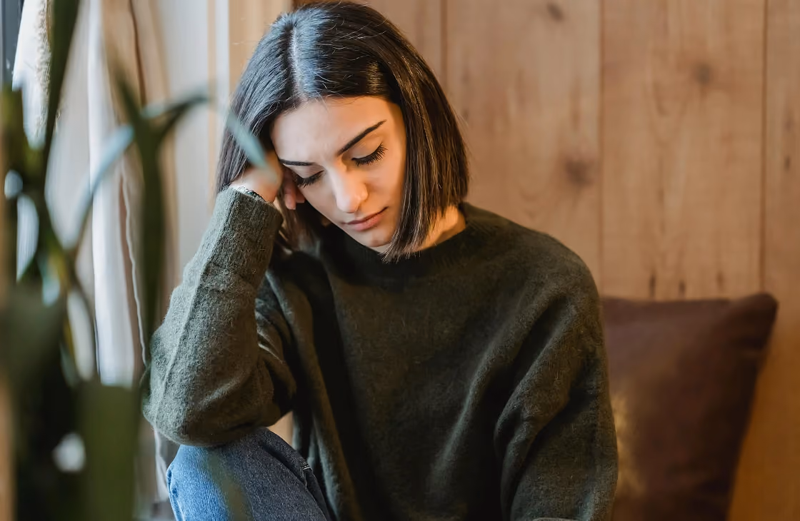 Young woman sitting indoors with her head resting on her hand, looking down thoughtfully.
