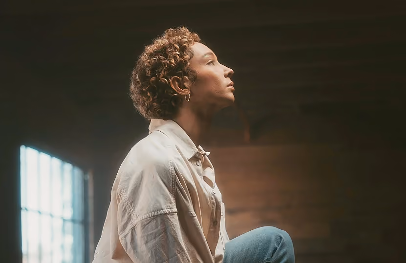 Side profile of a person with curly hair looking upward, wearing a white shirt and blue jeans in a dimly lit room with a window.