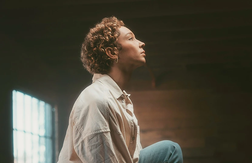 Side profile of a person with curly hair looking upward, wearing a white shirt and blue jeans in a dimly lit room with a window.