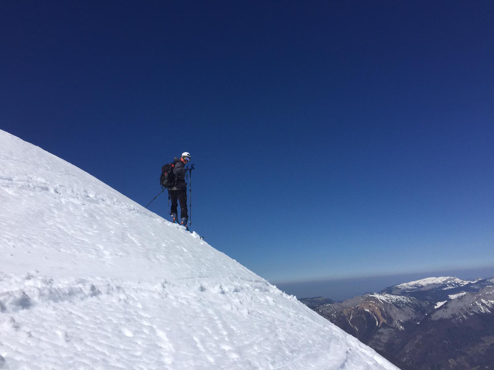 Uli Ebensperger, founder of Ziggma, pauses on a steep snow-covered ridge during a ski tour, looking back toward the camera. Distant alpine peaks and a deep blue sky stretch to the horizon.