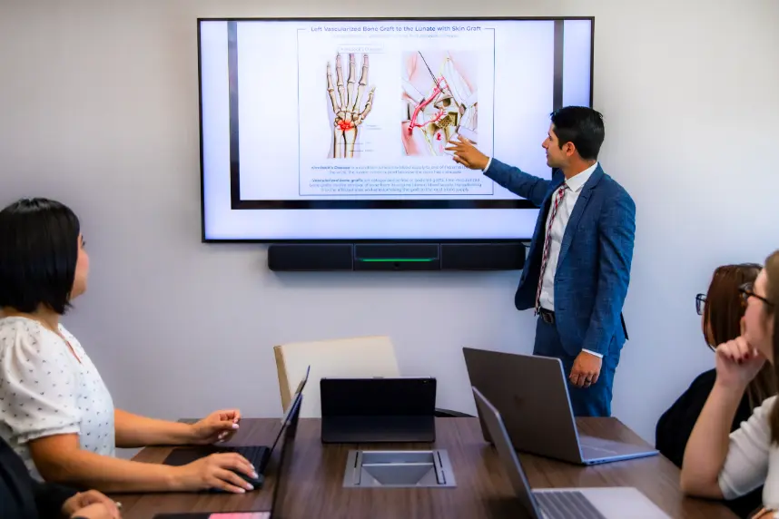 Professional delivering a medical presentation in a meeting, pointing to an anatomy diagram of a hand and a bone graft procedure shown on a large screen.