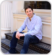 Smiling man in a light blue shirt and dark jeans sitting on outdoor metal stairs.