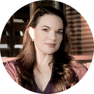 Portrait of a woman with long brown hair wearing a purple top, sitting indoors with wooden blinds in the background.