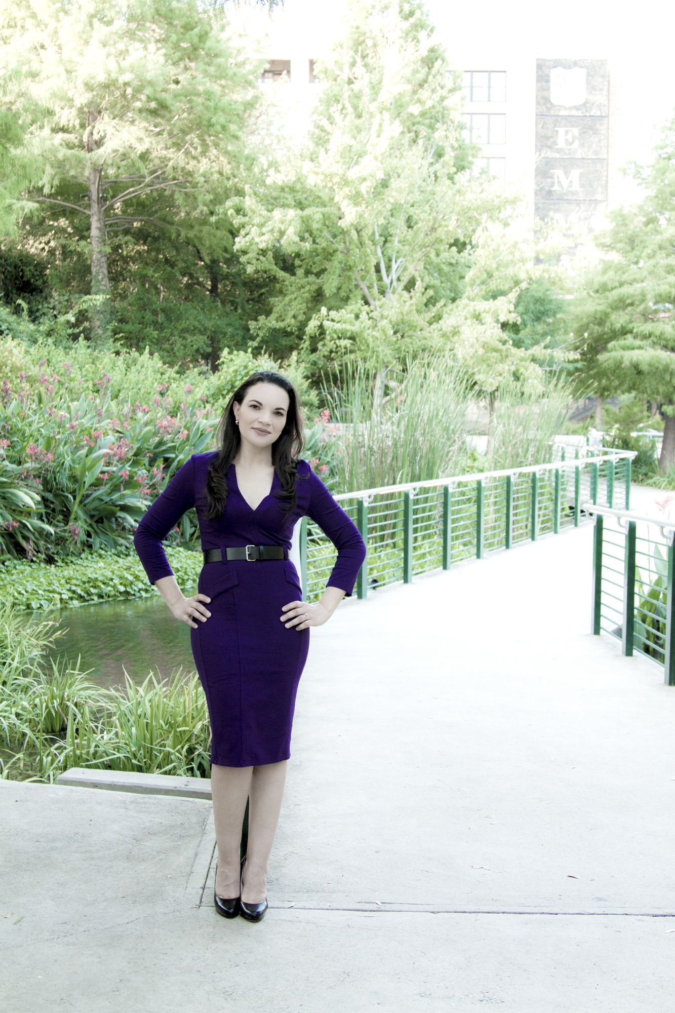 Woman wearing a fitted purple dress and black belt standing on a concrete path with greenery and flowers in the background.
