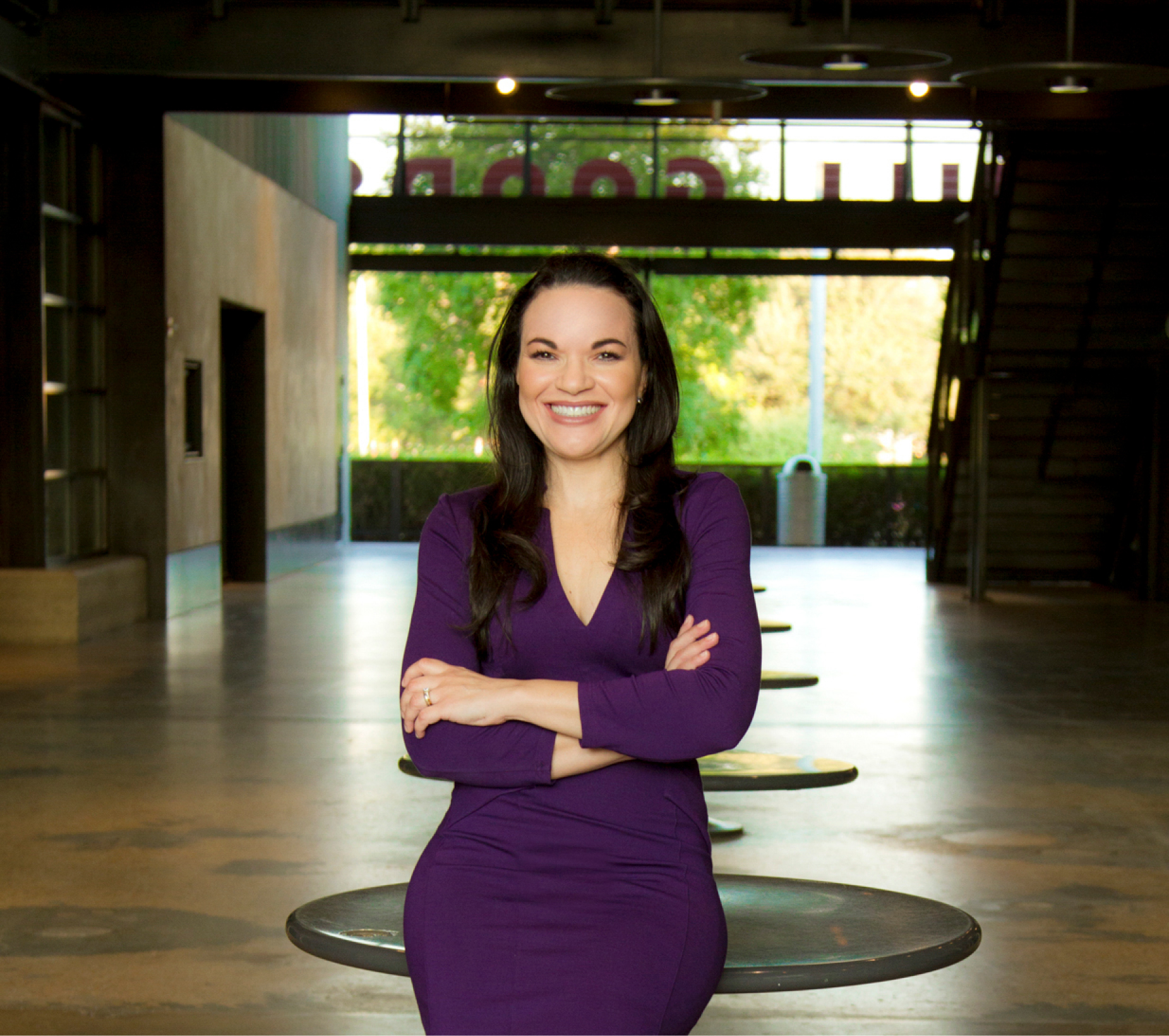 Smiling woman with long dark hair wearing a purple dress standing with arms crossed in a spacious indoor area.