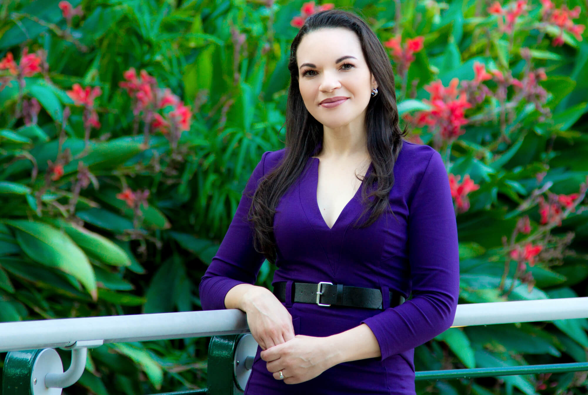 Woman with long dark hair wearing a purple dress and black belt, standing in front of green foliage with red flowers, leaning on a white railing.