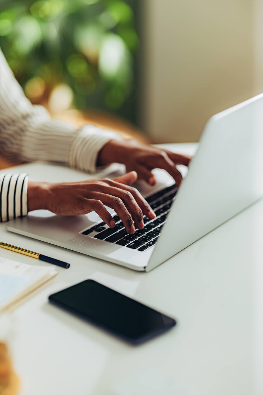 Person typing on a laptop keyboard at a white desk with a smartphone and pen nearby.