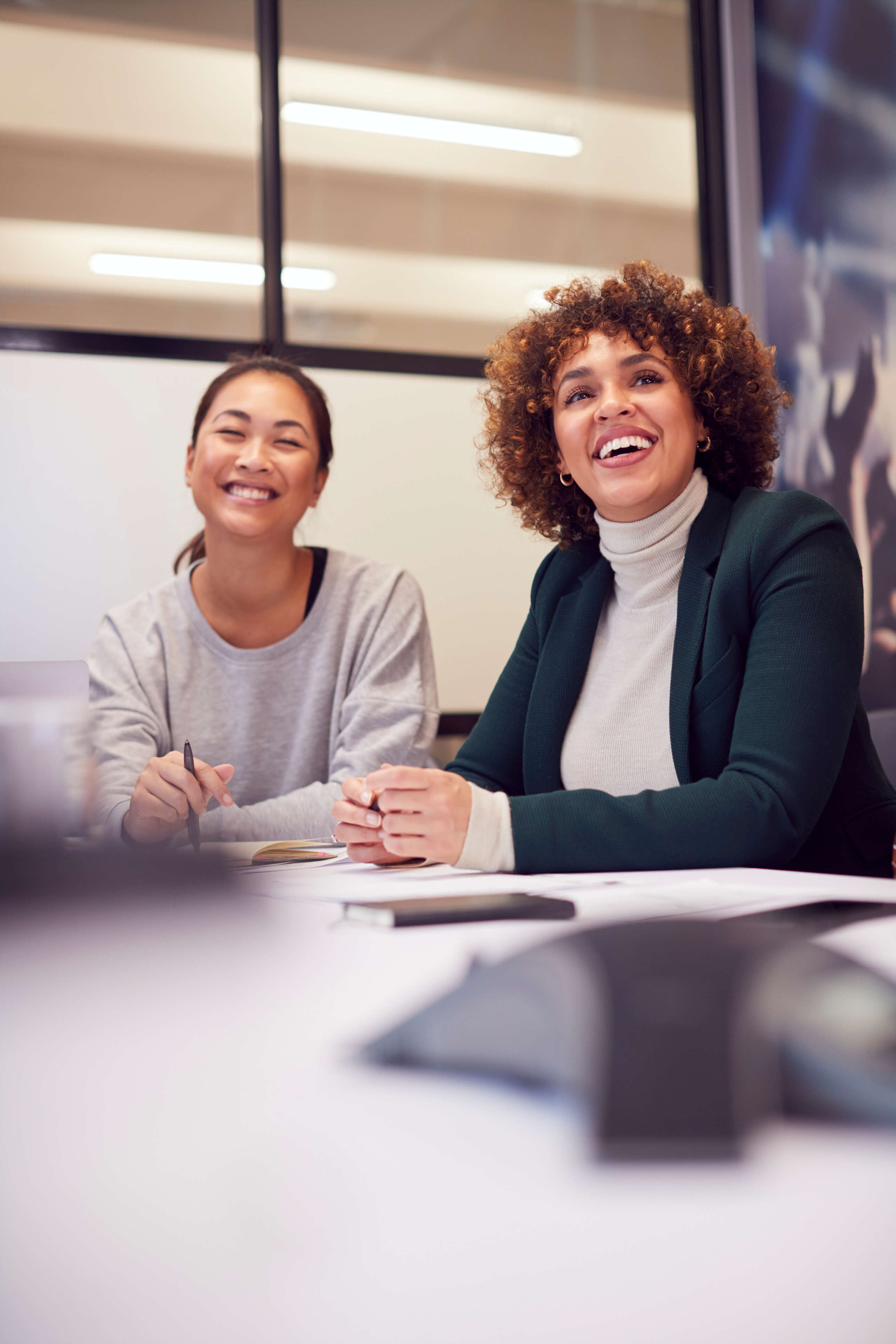 Two smiling women sitting at a table in an office, one holding a pen and the other clasping her hands.