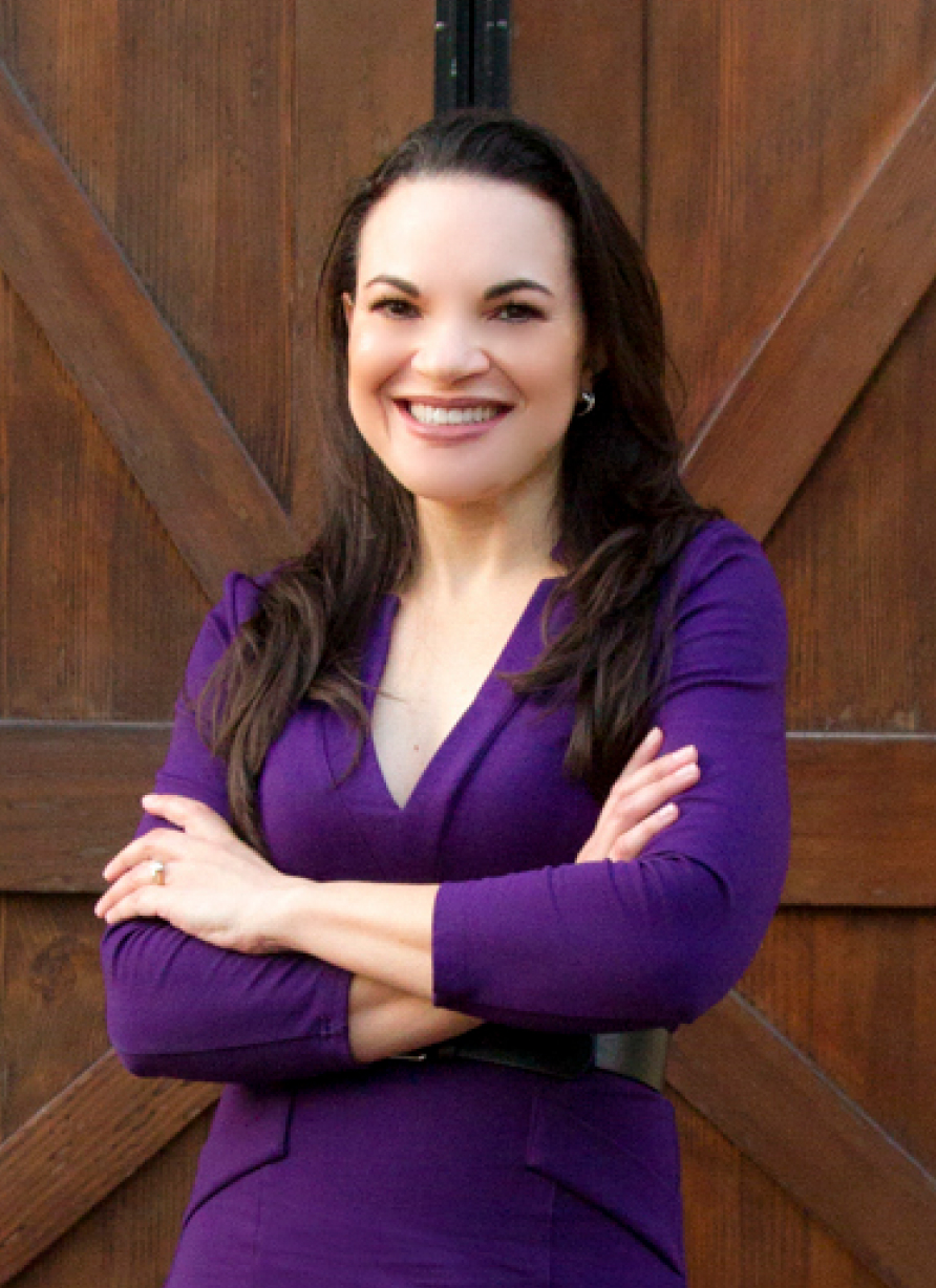 Smiling woman with long dark hair in a purple dress standing with arms crossed in front of wooden doors.