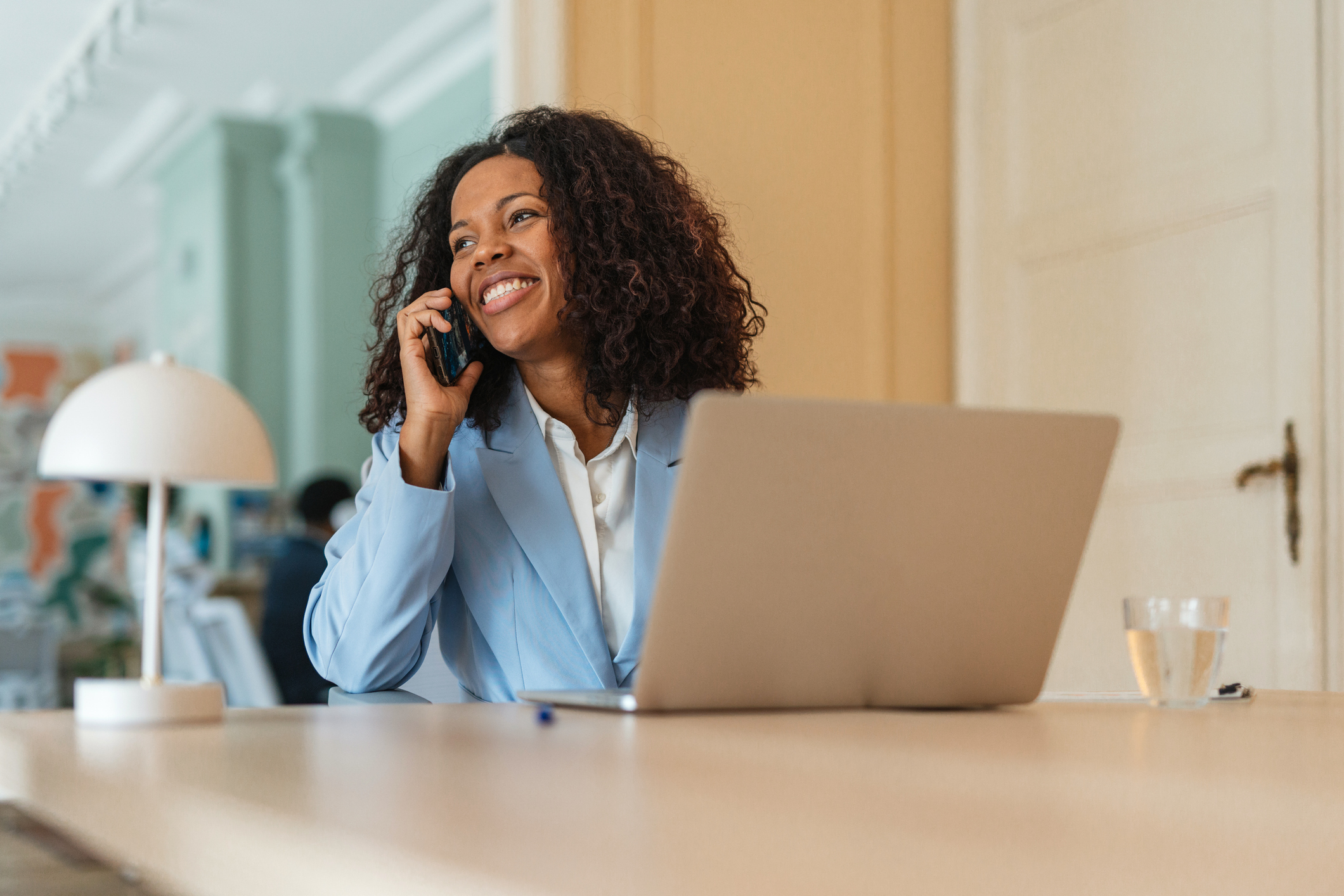 Smiling woman in light blue blazer talking on phone at a desk with laptop and lamp in a modern office.