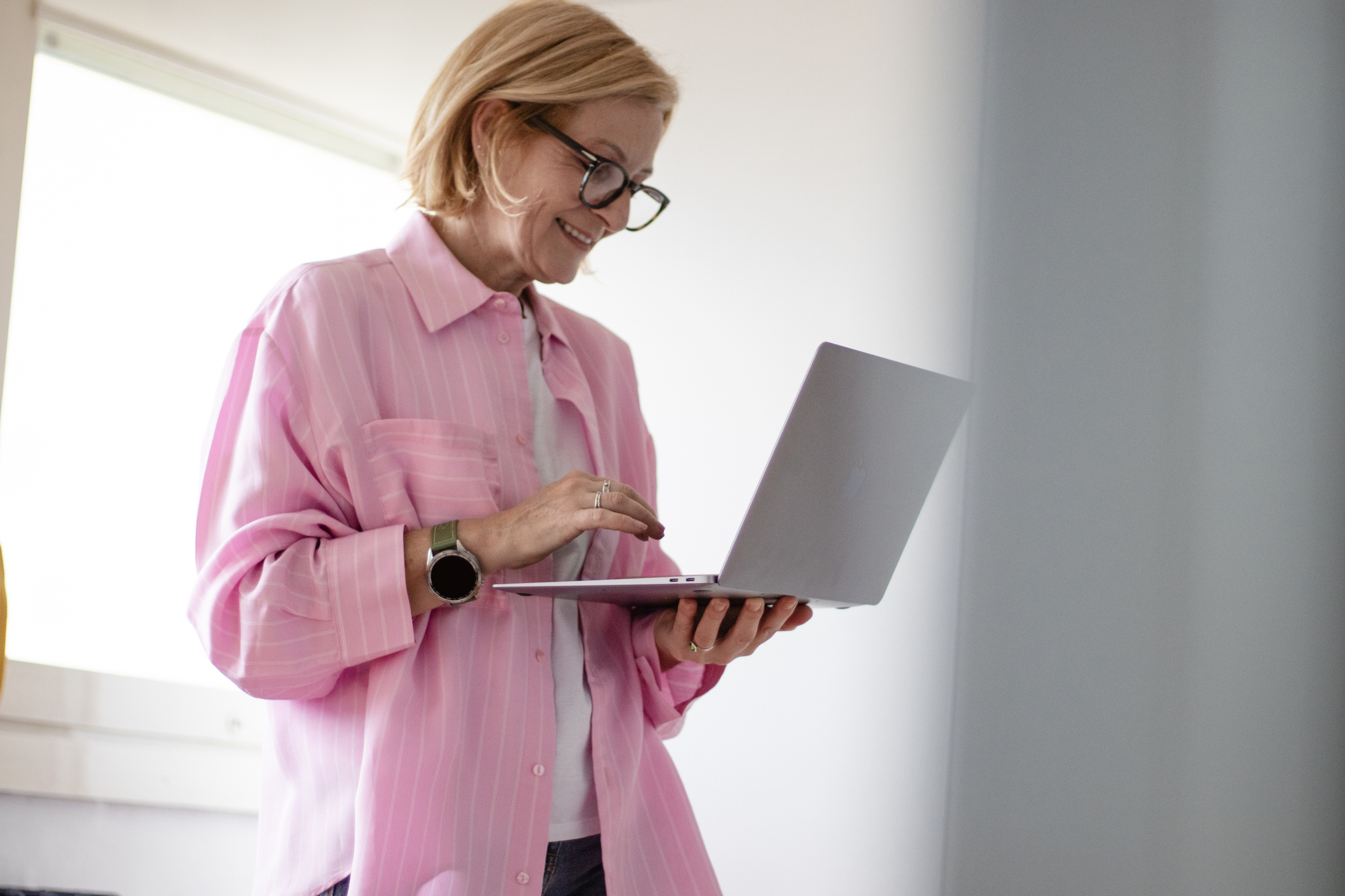 Smiling woman in glasses and pink shirt using a laptop indoors near a bright window.