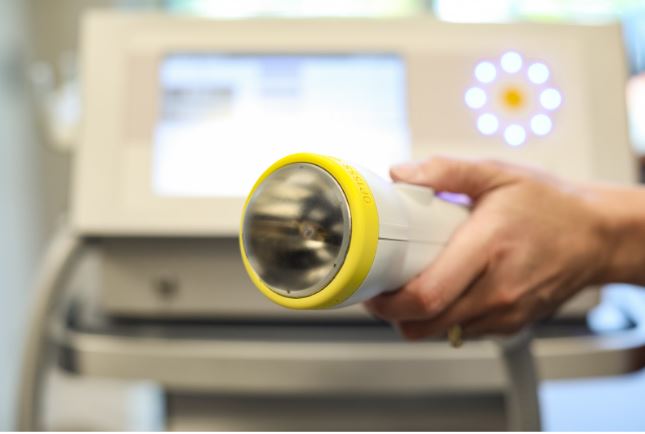 Close-up of a hand holding a white and yellow softwave therapy machine device with a blurred background.