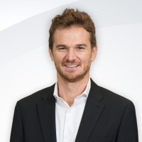 Hombre con traje negro y camisa blanca sonriendo frente a un fondo blanco claro.