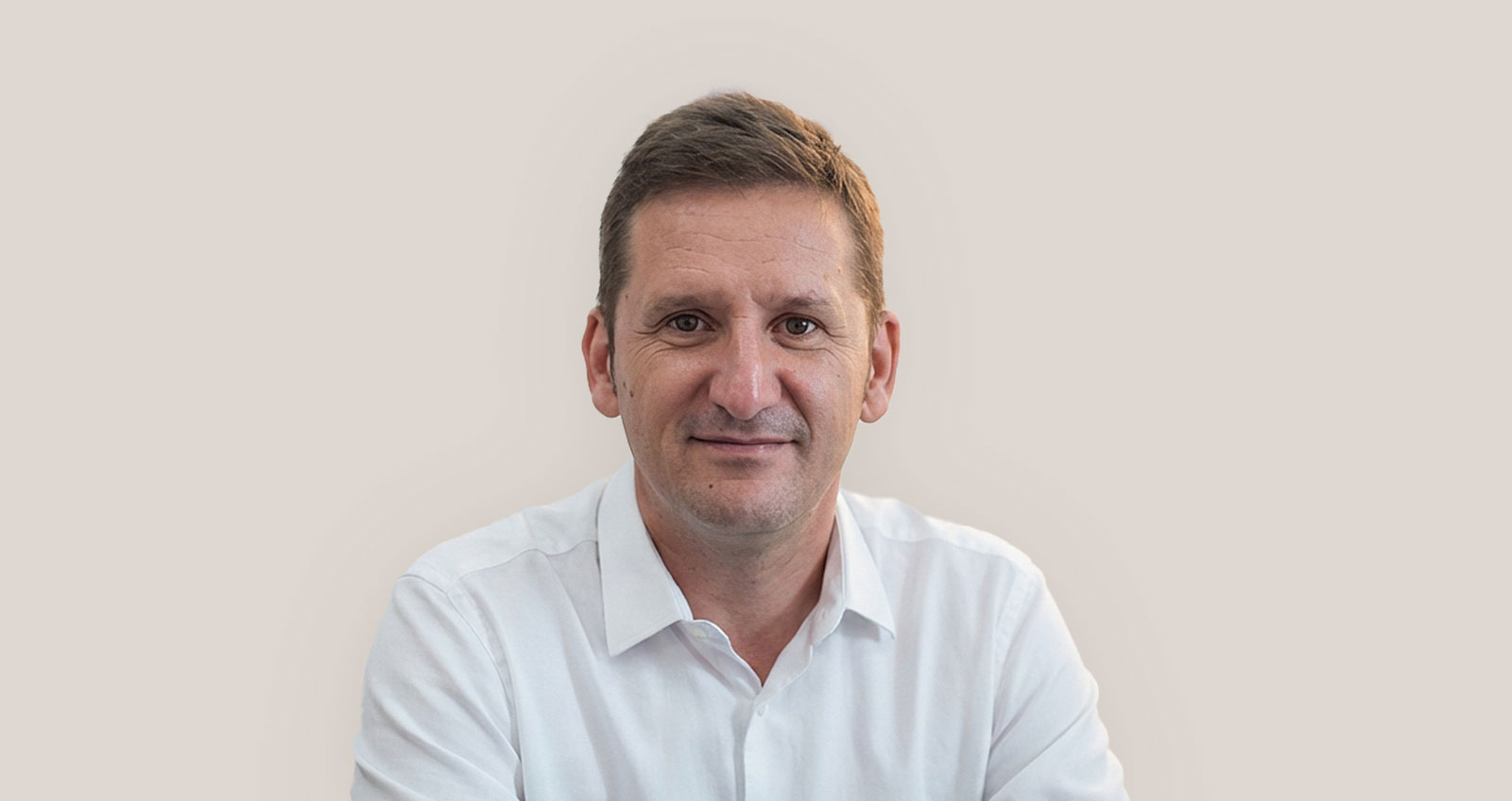 Hombre de mediana edad con camisa blanca sonriendo frente a fondo beige neutro.