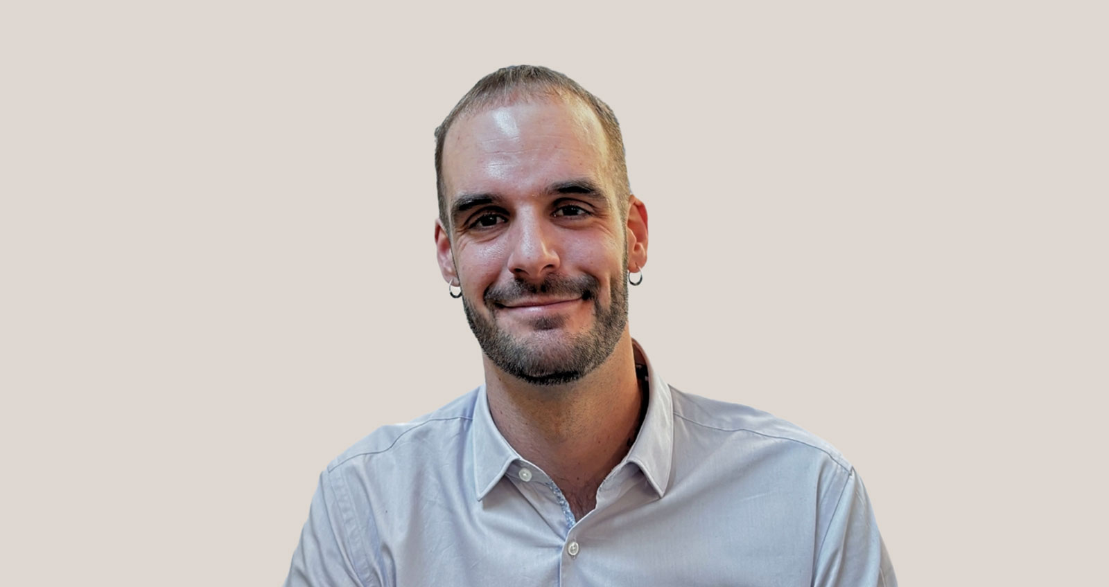Hombre con camisa clara, barba corta y pendientes en ambas orejas sonriendo frente a fondo neutro.