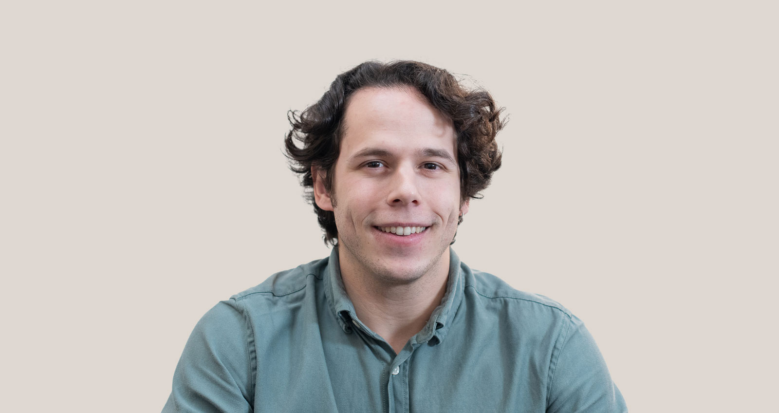 Joven con cabello rizado y camisa verde sonriente mirando a la cámara sobre un fondo claro.