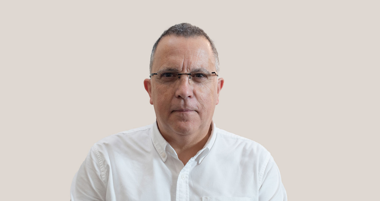 Hombre de mediana edad con gafas y camisa blanca, mirando directamente a la cámara sobre fondo beige.
