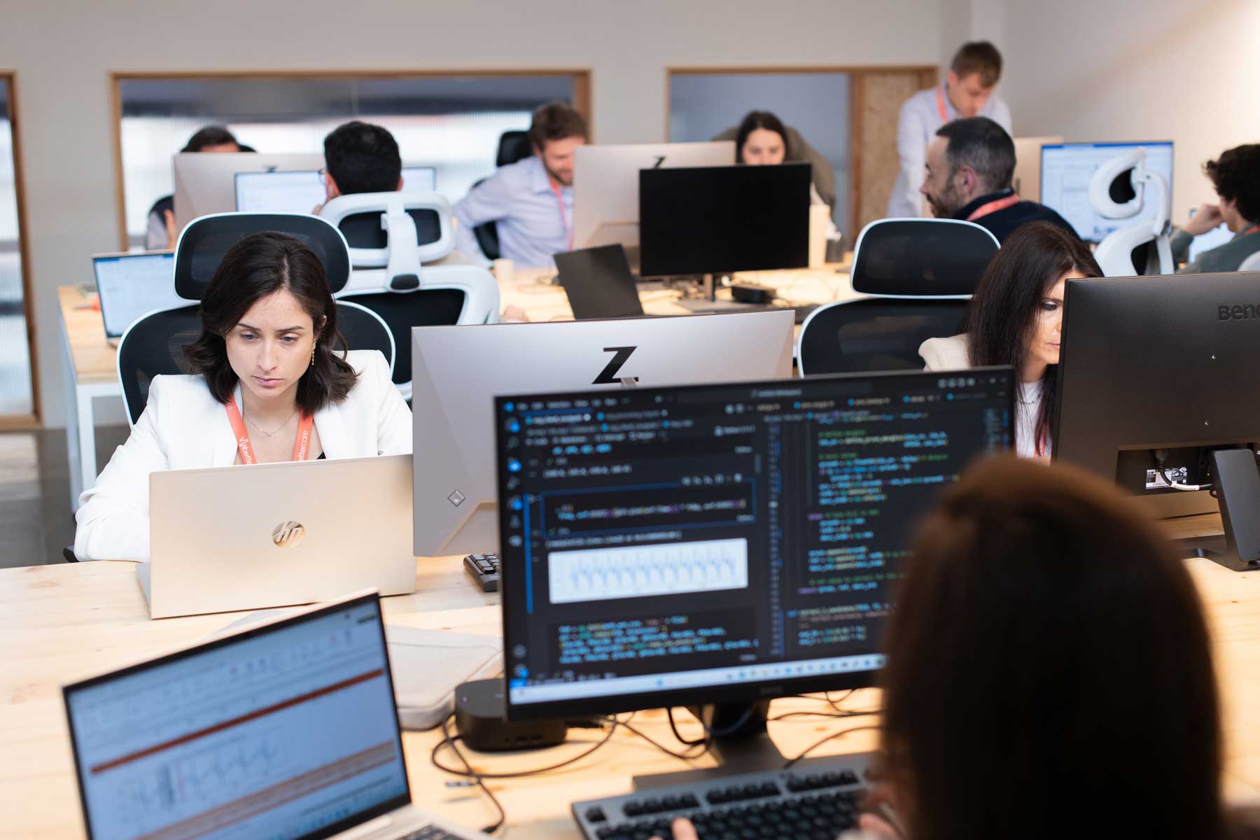 Varias personas trabajando en una oficina moderna con múltiples monitores y laptops, algunas con código en pantalla.