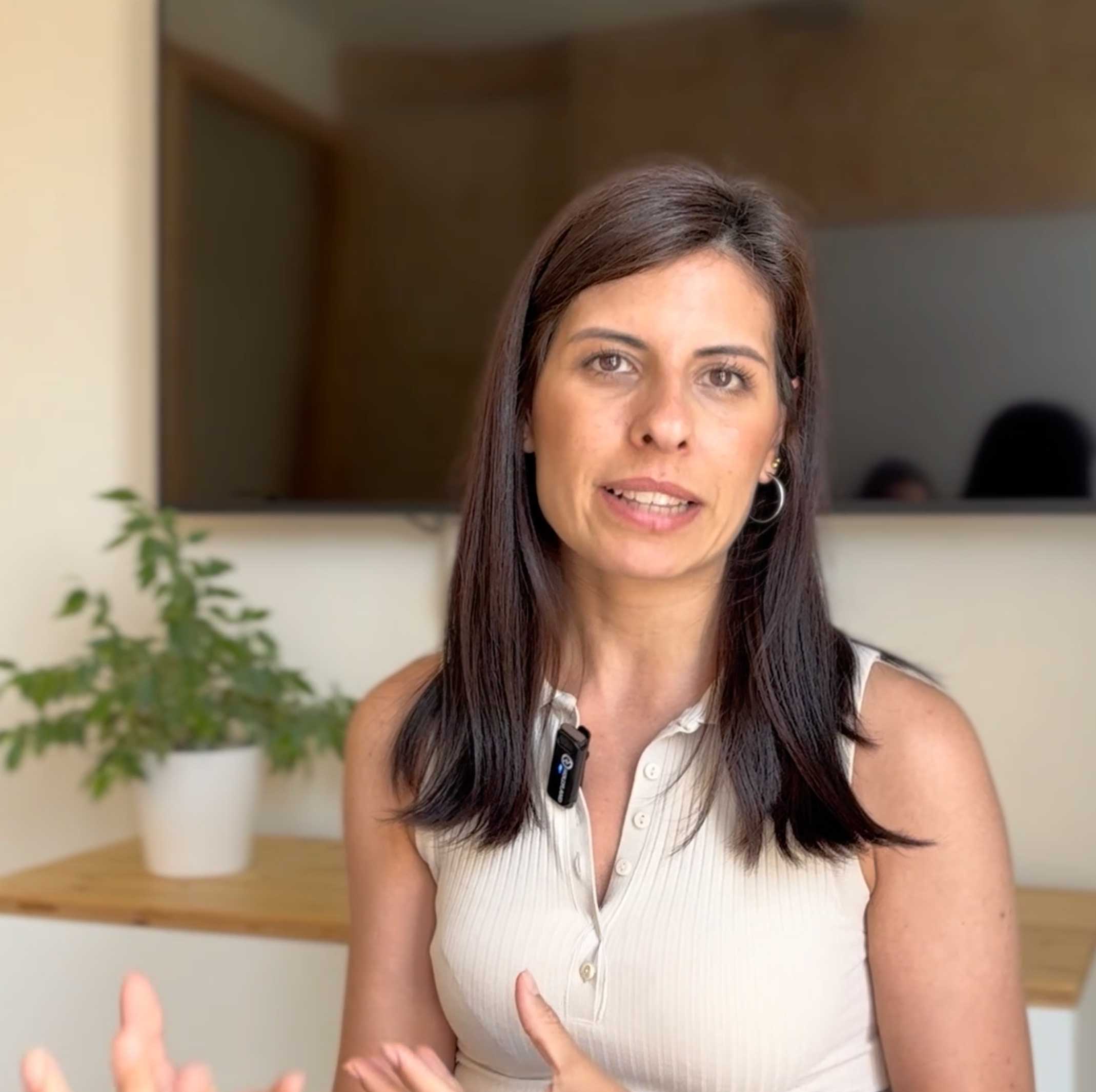 Mujer con cabello largo y oscuro hablando frente a una cámara, con una planta en maceta y una pantalla negra desenfocadas en el fondo.