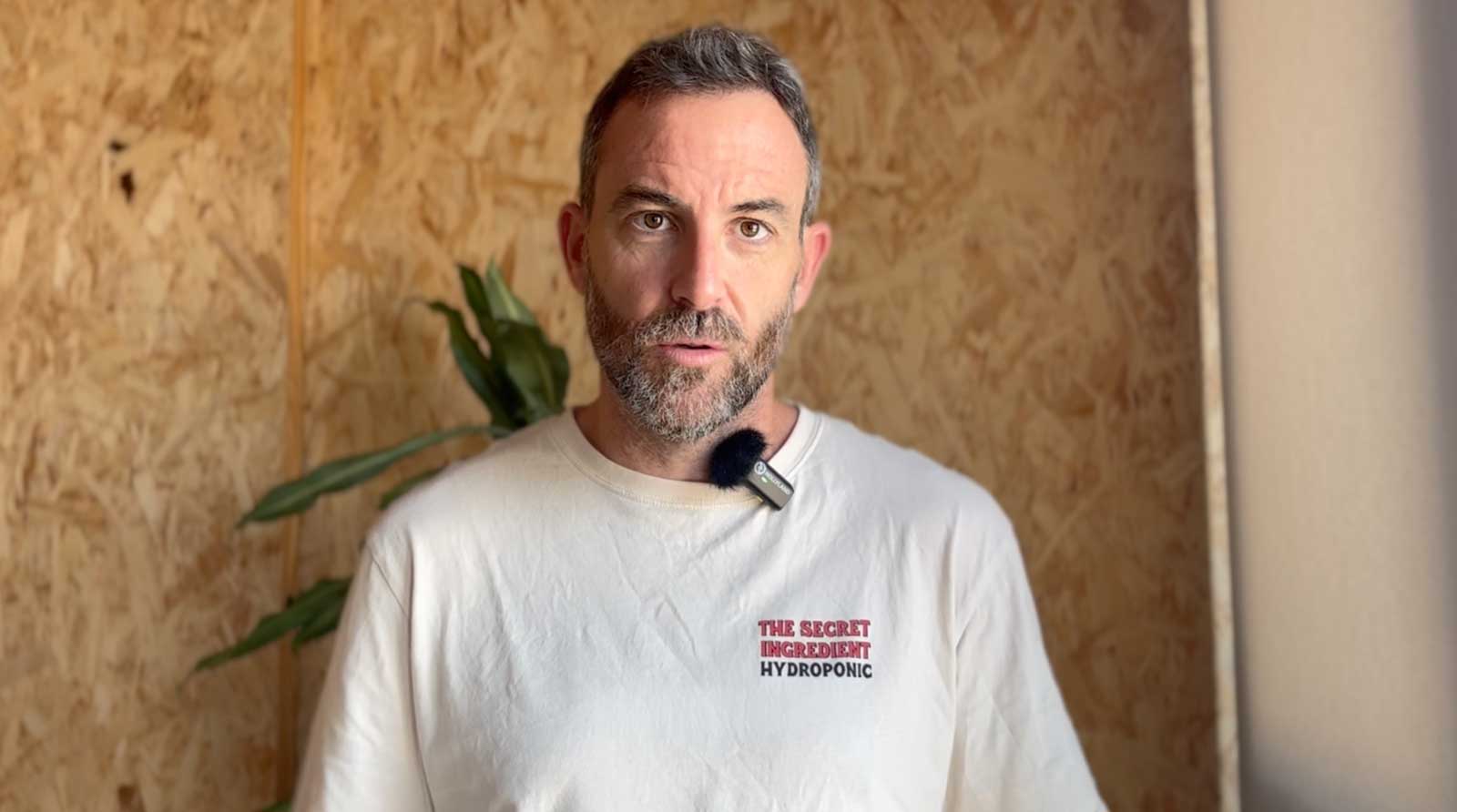 Hombre con barba y camiseta blanca con texto 'THE SECRET INGREDIENT HYDROPONIC' frente a una pared de madera con planta al fondo.