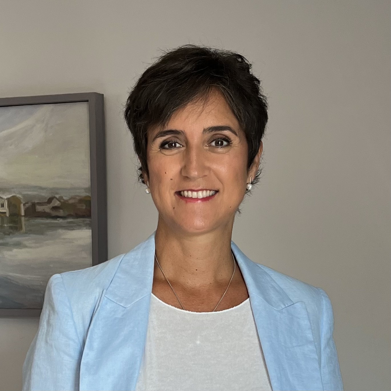 Mujer con cabello corto oscuro, chaqueta azul clara y camiseta blanca sonriente frente a un fondo neutro con un cuadro.