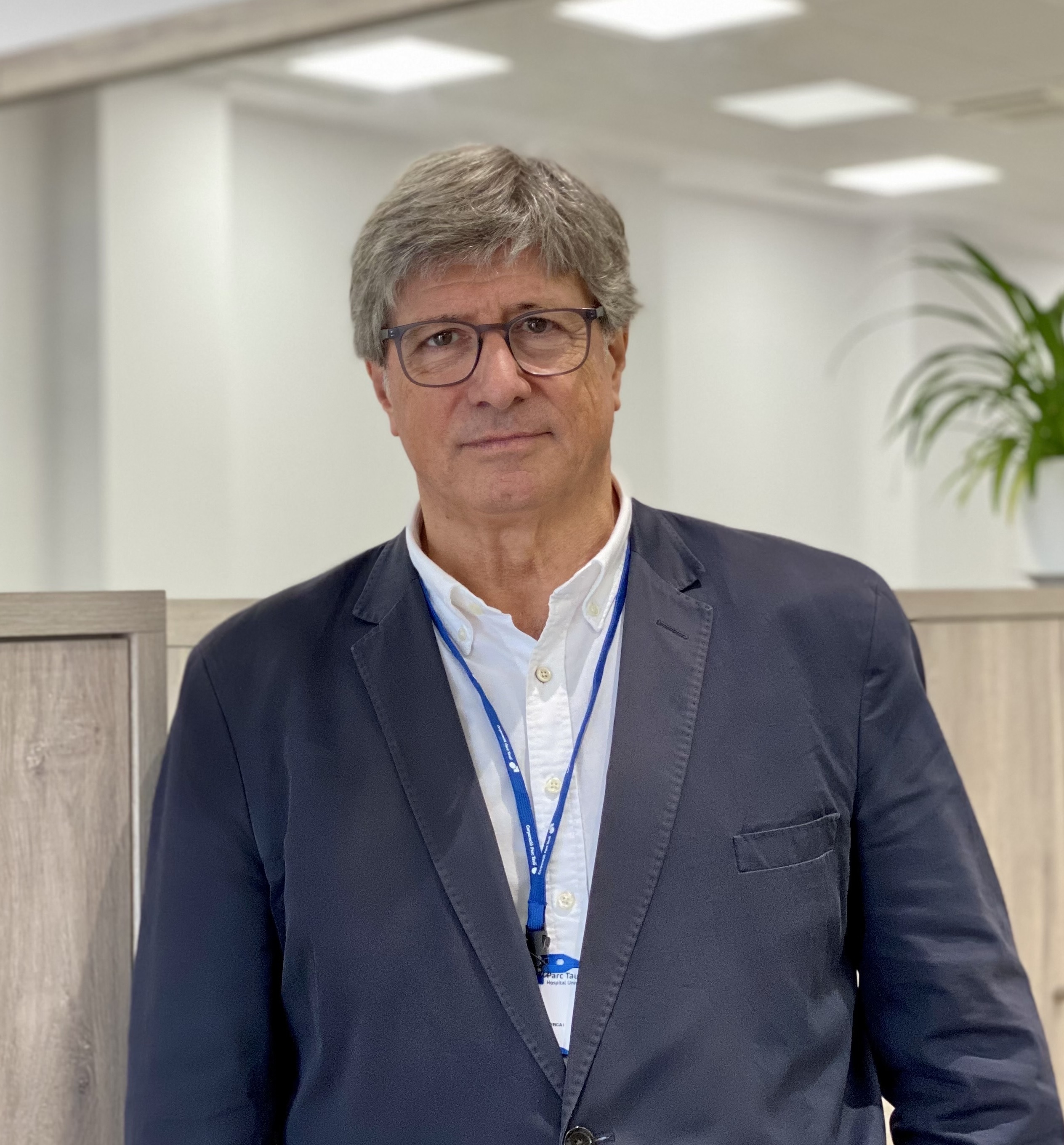Hombre de mediana edad con gafas, chaqueta azul y camisa blanca, con gafete azul, en una oficina moderna.