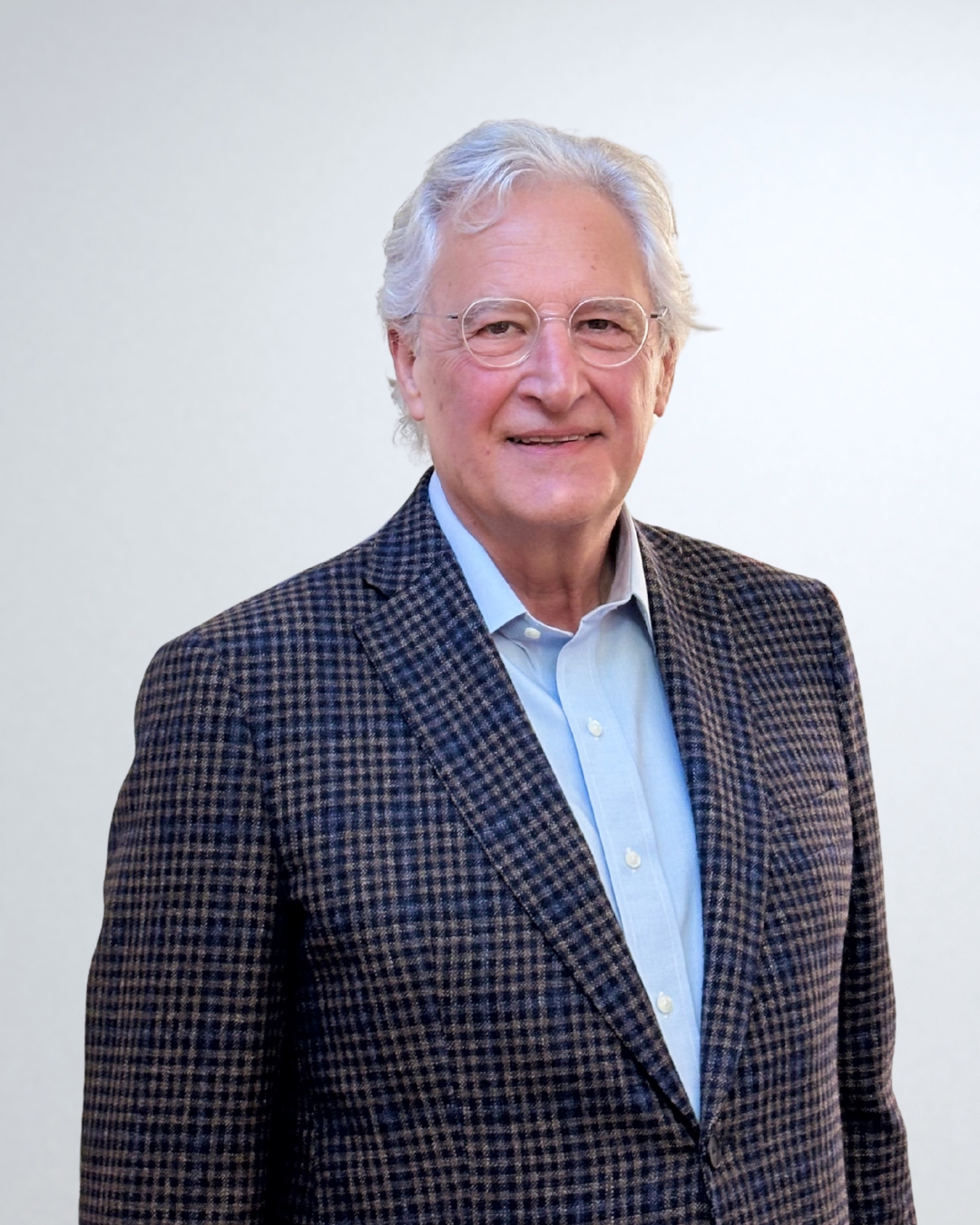 Hombre mayor con cabello canoso y gafas, vestido con camisa azul clara y chaqueta de cuadros marrón y azul.